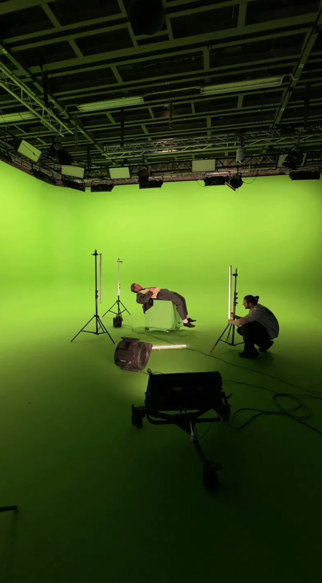 Behind the scenes on a green screen film shoot with a model on a cube, lighting stands, and crew at work